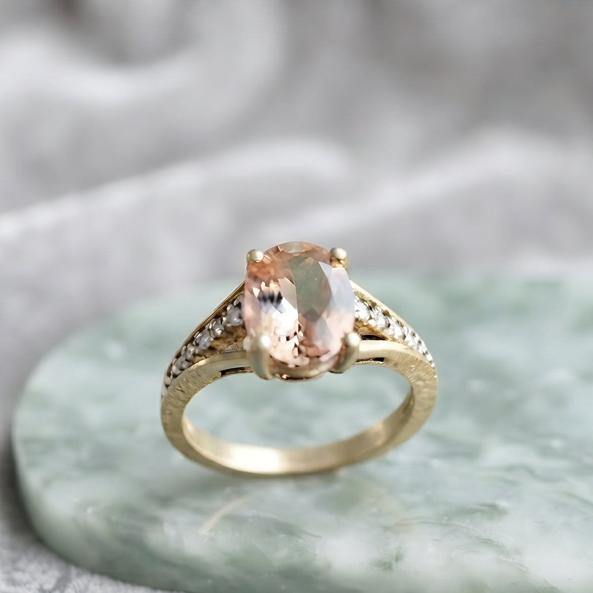 A gold ring with a pear-cut peach-colored gemstone and small accent stones on the band, displayed on a light green marble slab against a soft gray fabric background.