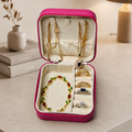 A pink jewelry organizer box with a cream interior, shown open on a tabletop. Inside and on the lid are several gold-tone jewelry pieces, including necklaces and rings, arranged in compartments.