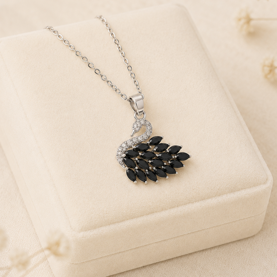 A silver-toned necklace with a peacock pendant, featuring black marquise-cut stones and clear crystal accents, displayed on a cream-colored velvet jewelry box with dried flowers in the background.