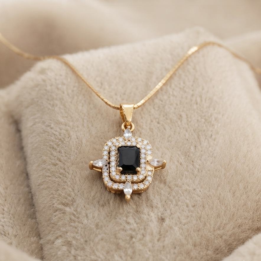 A gold pendant necklace with a black rectangular center stone and surrounding clear stones, displayed on a soft beige fabric surface.