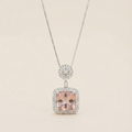 A silver-toned necklace featuring a square pendant with a central blush pink stone, surrounded by a halo of small clear stones, suspended from a delicate silver chain against a light beige background.