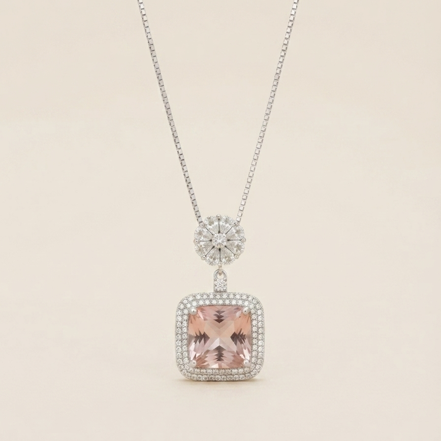 A silver-toned necklace featuring a square pendant with a central blush pink stone, surrounded by a halo of small clear stones, suspended from a delicate silver chain against a light beige background.