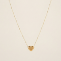 A delicate gold chain necklace featuring a small, polished gold heart pendant, displayed against a solid, warm off-white background.