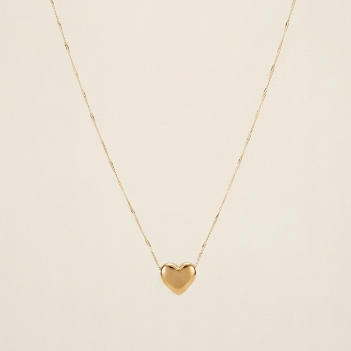 A delicate gold chain necklace featuring a small, polished gold heart pendant, displayed against a solid, warm off-white background.
