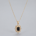 A gold pendant necklace featuring a rectangular black gemstone surrounded by a halo of small clear stones, suspended from a delicate gold chain against a light gray background.