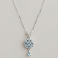 A silver-toned pendant necklace featuring a circular pavé design with light blue gemstones and a dangling teardrop-shaped light blue stone, shown against a plain light beige background.