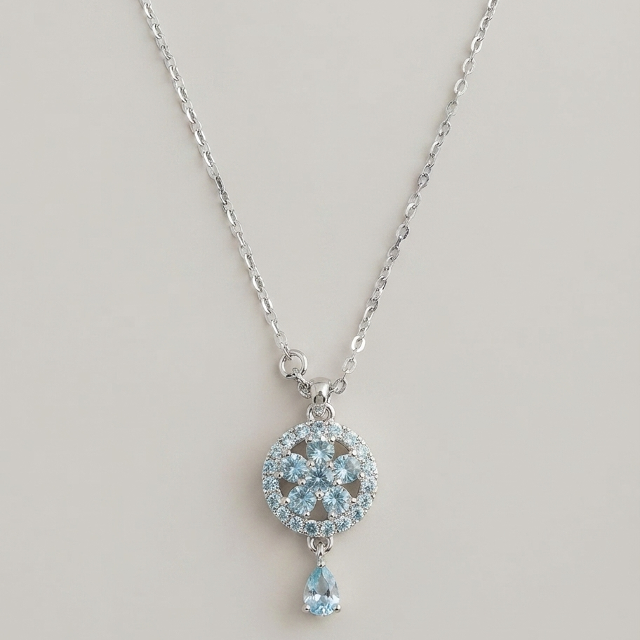A silver-toned pendant necklace featuring a circular pavé design with light blue gemstones and a dangling teardrop-shaped light blue stone, shown against a plain light beige background.