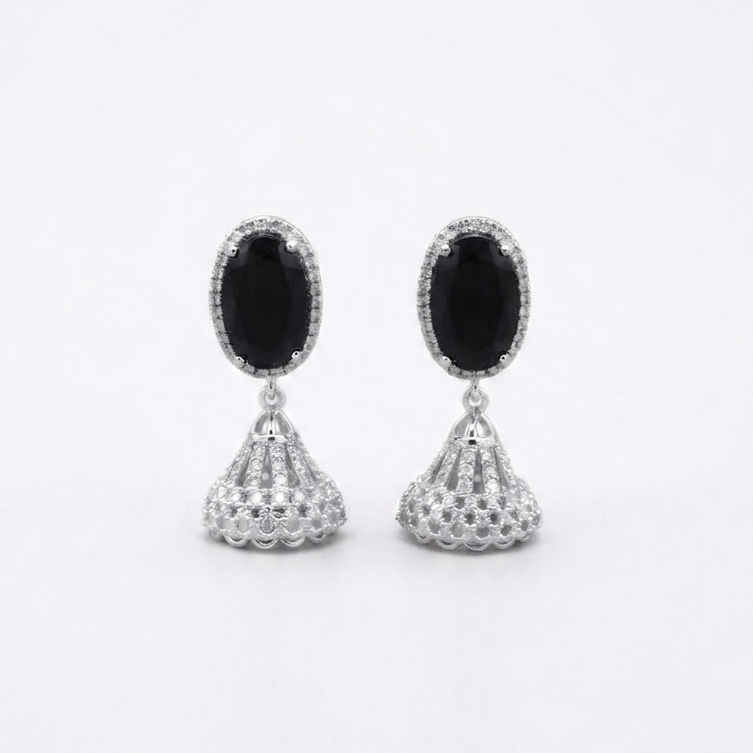 A pair of silver-toned dangle earrings with oval black stones and textured triangular drops, displayed on a white fabric background with warm fairy lights.
