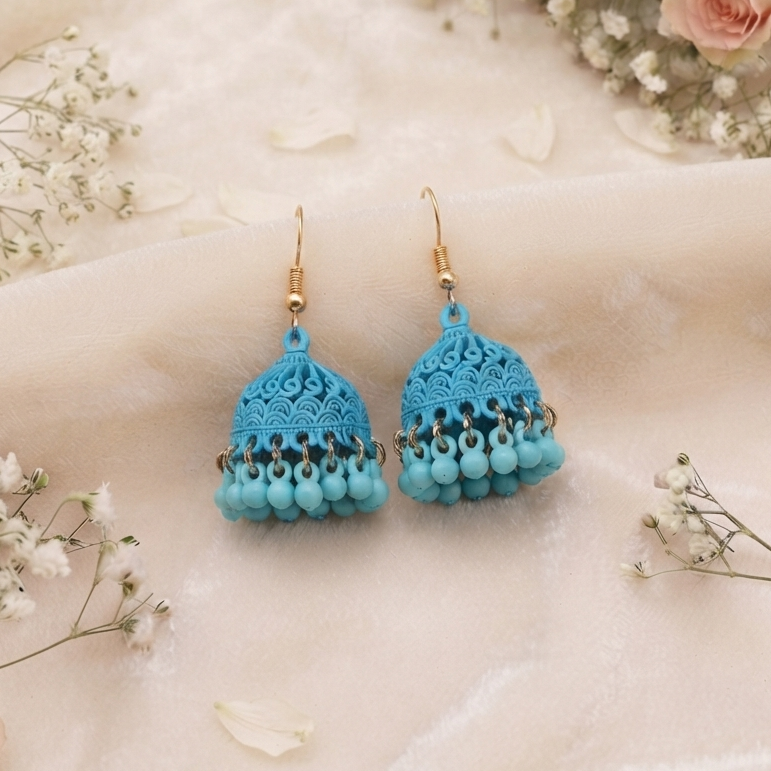 A pair of turquoise blue crochet jhumka earrings with gold-tone hooks, displayed on a soft beige fabric backdrop decorated with pink roses and white baby's breath. The brand logo 'ZIVARO' is visible in the top left, and the website 'www.