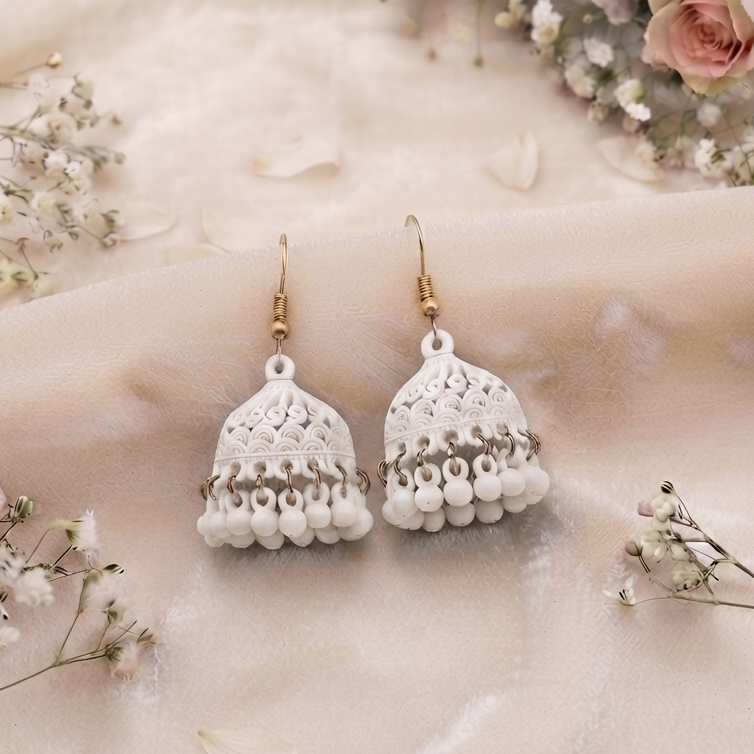 A pair of turquoise blue crochet jhumka earrings with gold-tone hooks, displayed on a soft beige fabric backdrop decorated with pink roses and white baby's breath. The brand logo 'ZIVARO' is visible in the top left, and the website 'www.