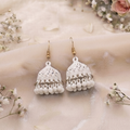 A pair of white embroidered jhumka earrings with gold-tone hooks, displayed on a soft beige fabric backdrop with pink roses and baby's breath flowers. The brand name ZIVARO is visible at the top left, and the website www.