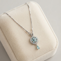A silver-tone necklace with a floral cluster pendant and a teardrop charm in light blue, displayed on a cream-colored velvet jewelry box.