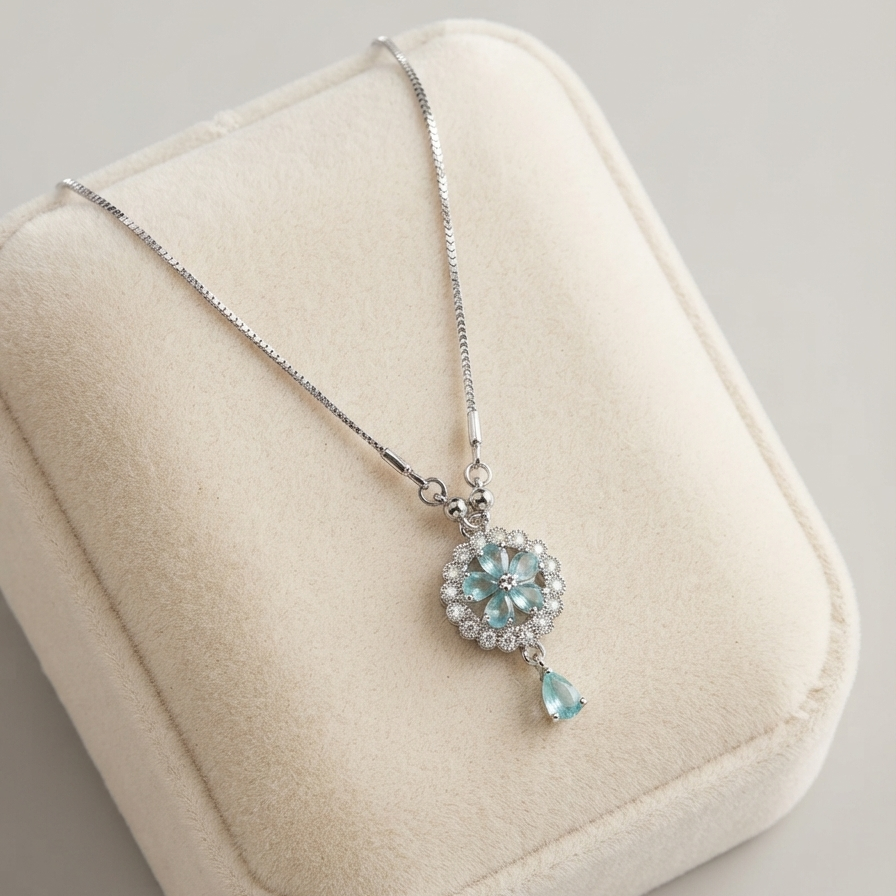 A silver-tone necklace with a floral cluster pendant and a teardrop charm in light blue, displayed on a cream-colored velvet jewelry box.