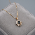 A gold pendant necklace with a central black rectangular stone and surrounding clear stones, displayed on a gray velvet jewelry box.