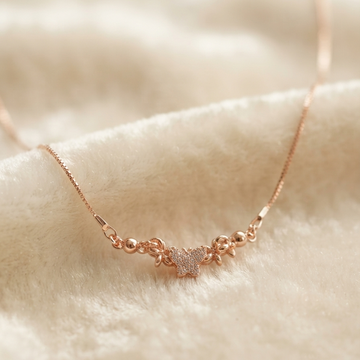 A delicate rose gold necklace with a small central charm, resting on a soft, textured cream-colored fabric background.