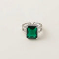 A silver-toned split-band ring featuring a rectangular emerald-cut green gemstone surrounded by a halo of small clear stones, set against a plain off-white background.