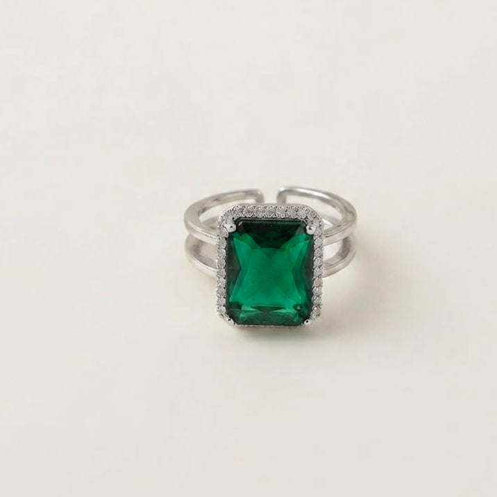 A silver-toned split-band ring featuring a rectangular emerald-cut green gemstone surrounded by a halo of small clear stones, set against a plain off-white background.