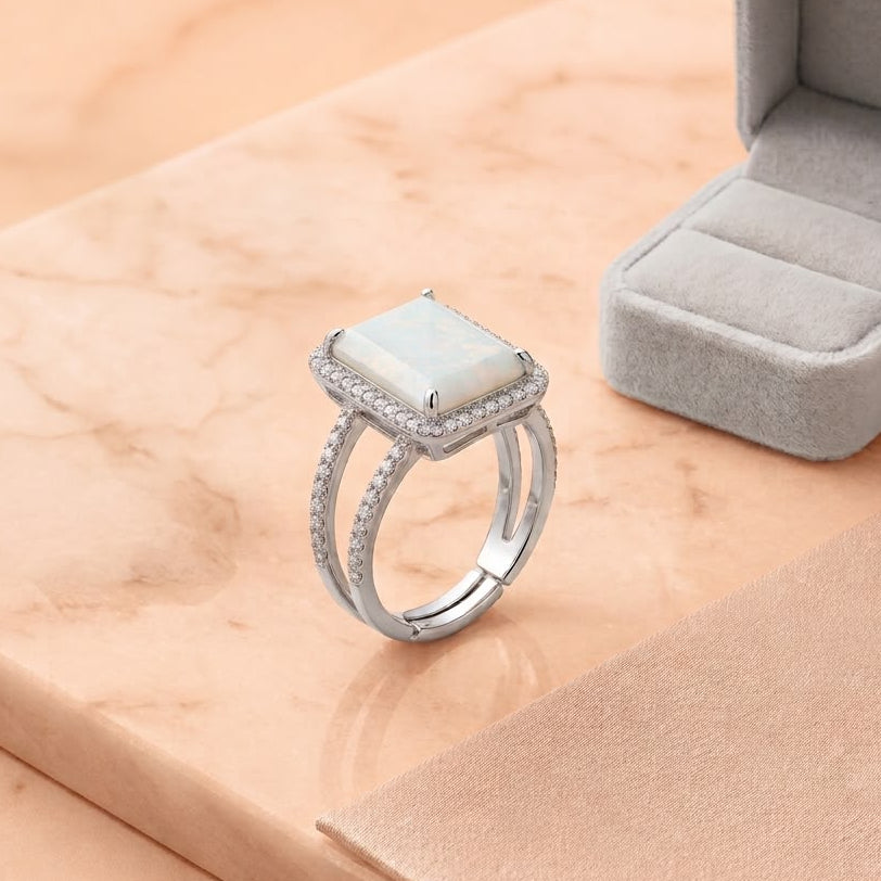 A silver ring with a rectangular white stone and small clear accent stones, displayed on a peach marble surface next to an open gray ring box. A Shopify Product watermark is visible at the bottom.