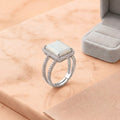 A silver ring with a rectangular white stone and small clear accent stones, displayed on a peach marble surface next to an open gray ring box. A Shopify Product watermark is visible at the bottom.