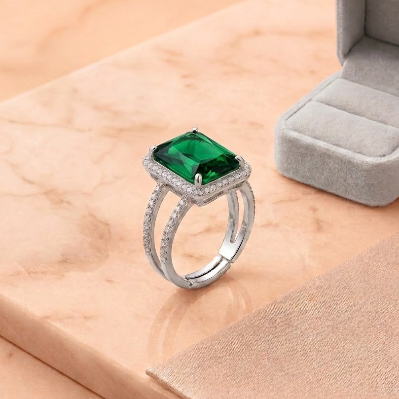 A silver ring with a square-cut green gemstone and small clear stones in a halo setting, displayed on a peach marble surface next to an open gray ring box.