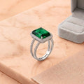 A silver ring with a square-cut green gemstone and small clear stones in a halo setting, displayed on a peach marble surface next to an open gray ring box.