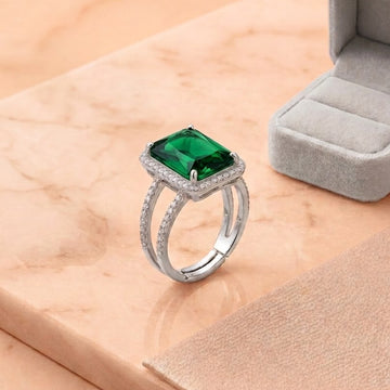 A silver ring with a square-cut green gemstone and small clear stones in a halo setting, displayed on a peach marble surface next to an open gray ring box.