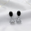 A pair of silver-toned dangle earrings with oval black stones and textured triangular drops, displayed on a white fabric background with warm fairy lights.