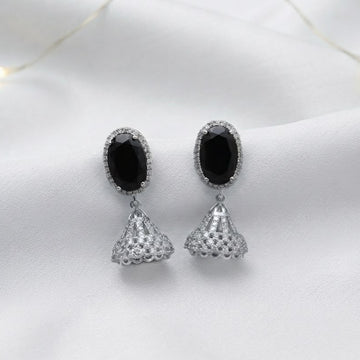A pair of silver-toned dangle earrings with oval black stones and textured triangular drops, displayed on a white fabric background with warm fairy lights.